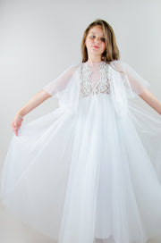 Floor-length white tulle princess dress with a floating tulle skirt, transparent butterfly sleeves and transparent bodice covered with embellished flower details and pearls. Girl dress for flower girls, weddings, communion or birthday party.