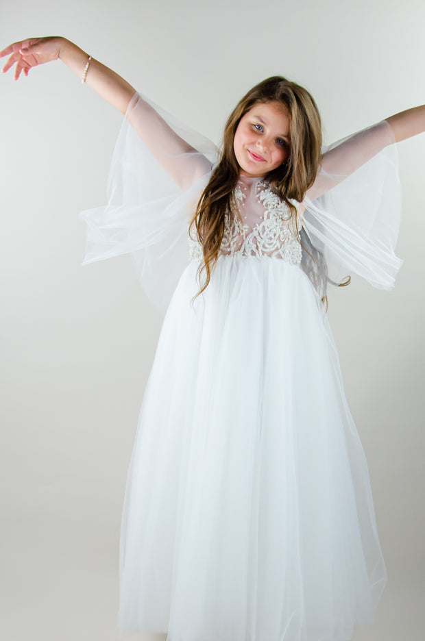 Floor-length white tulle princess dress with a floating tulle skirt, transparent butterfly sleeves and transparent bodice covered with embellished flower details and pearls. Girl dress for flower girls, weddings, communion or birthday party.