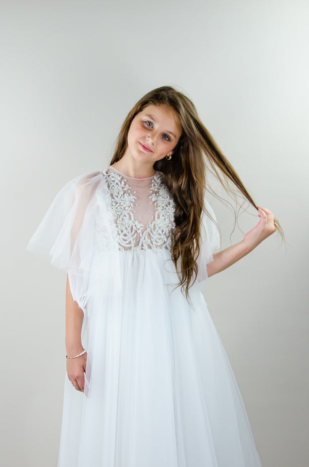 Floor-length white tulle princess dress with a floating tulle skirt, transparent butterfly sleeves and transparent bodice covered with embellished flower details and pearls. Girl dress for flower girls, weddings, communion or birthday party.