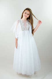 Floor-length white tulle princess dress with a floating tulle skirt, transparent butterfly sleeves and transparent bodice covered with embellished flower details and pearls. Girl dress for flower girls, weddings, communion or birthday party.