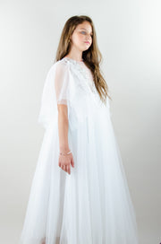 Floor-length white tulle princess dress with a floating tulle skirt, transparent butterfly sleeves and transparent bodice covered with embellished flower details and pearls. Girl dress for flower girls, weddings, communion or birthday party.