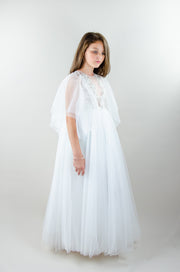 Floor-length white tulle princess dress with a floating tulle skirt, transparent butterfly sleeves and transparent bodice covered with embellished flower details and pearls. Girl dress for flower girls, weddings, communion or birthday party.