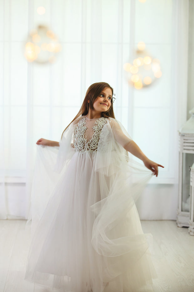 Floor-length white tulle princess dress with a floating tulle skirt, transparent butterfly sleeves and transparent bodice covered with embellished flower details and pearls. Girl dress for flower girls, weddings, communion or birthday party.