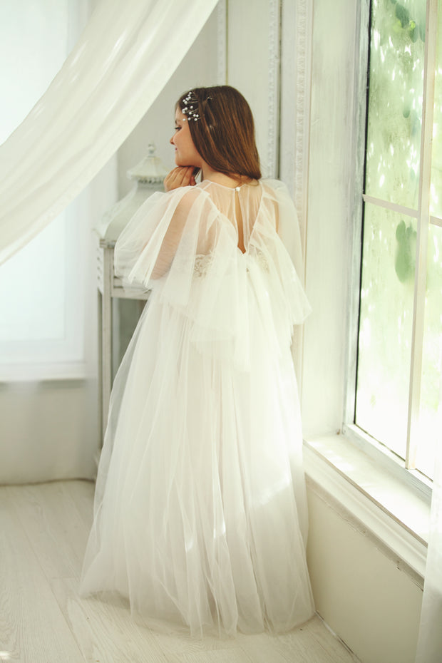 Floor-length white tulle princess dress with a floating tulle skirt, transparent butterfly sleeves and transparent bodice covered with embellished flower details and pearls. Girl dress for flower girls, weddings, communion or birthday party.