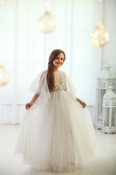 Floor-length white tulle princess dress with a floating tulle skirt, transparent butterfly sleeves and transparent bodice covered with embellished flower details and pearls. Girl dress for flower girls, weddings, communion or birthday party.