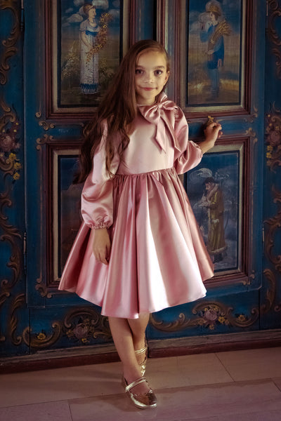 Knee-length satin girl dress in a blush pink colour. The dress features an a-line satin skirt with tulle underskirt which gives it volume. Satin top with high neckline is accentuated with a cute bow and long satin sleeves. For special occasions: Wedding, Birthday party, Prom, Flower girl, Eid and other events.