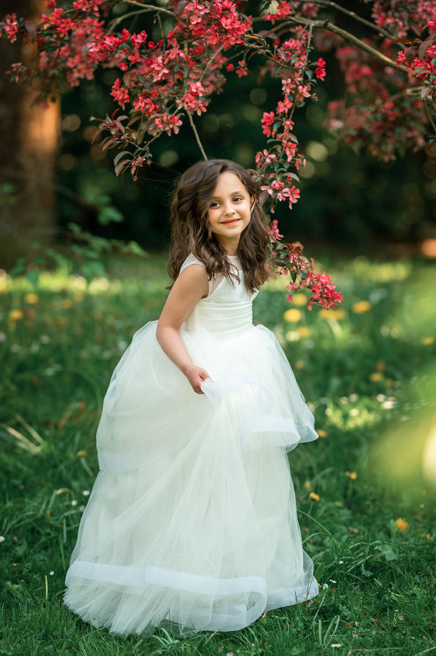 Long, white A-line flower girl dress with an asymmetrical multi-layer tulle skirt and white satin top with pleated satin details.