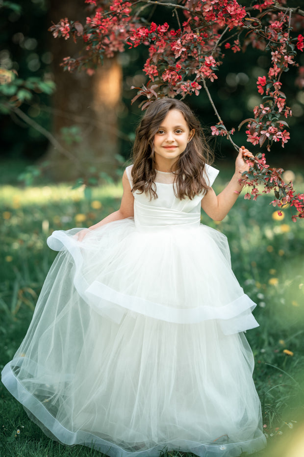 Long, white A-line flower girl dress with an asymmetrical multi-layer tulle skirt and white satin top with pleated satin details.