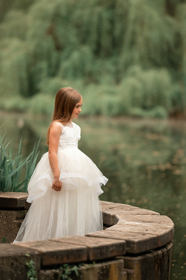Long, white A-line flower girl dress with an asymmetrical multi-layer tulle skirt and white satin top with pleated satin details.
